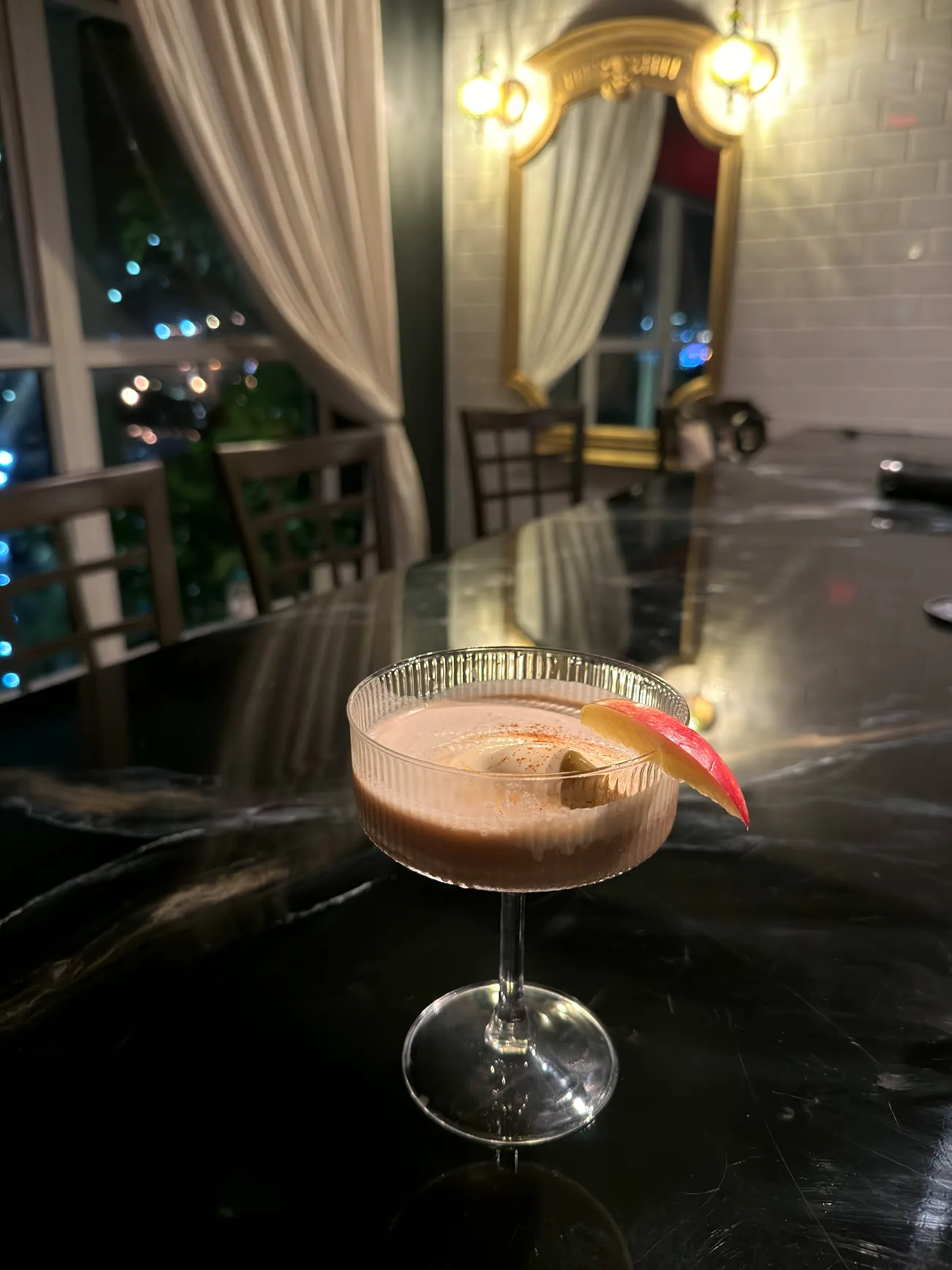 A ribbed coupe cocktail on the black marble bar at night, gold mirror and sconces glowing behind
