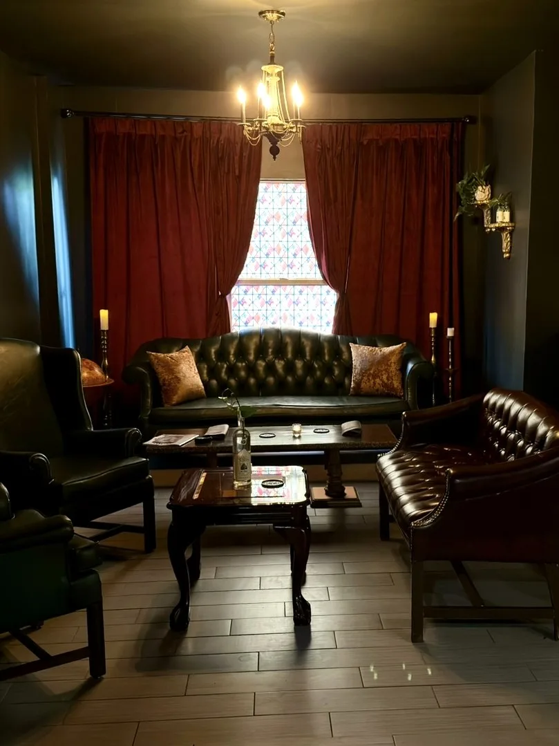 The parlor — leather chesterfield sofas around a carved coffee table, red velvet curtains and a stained-glass window under a brass chandelier